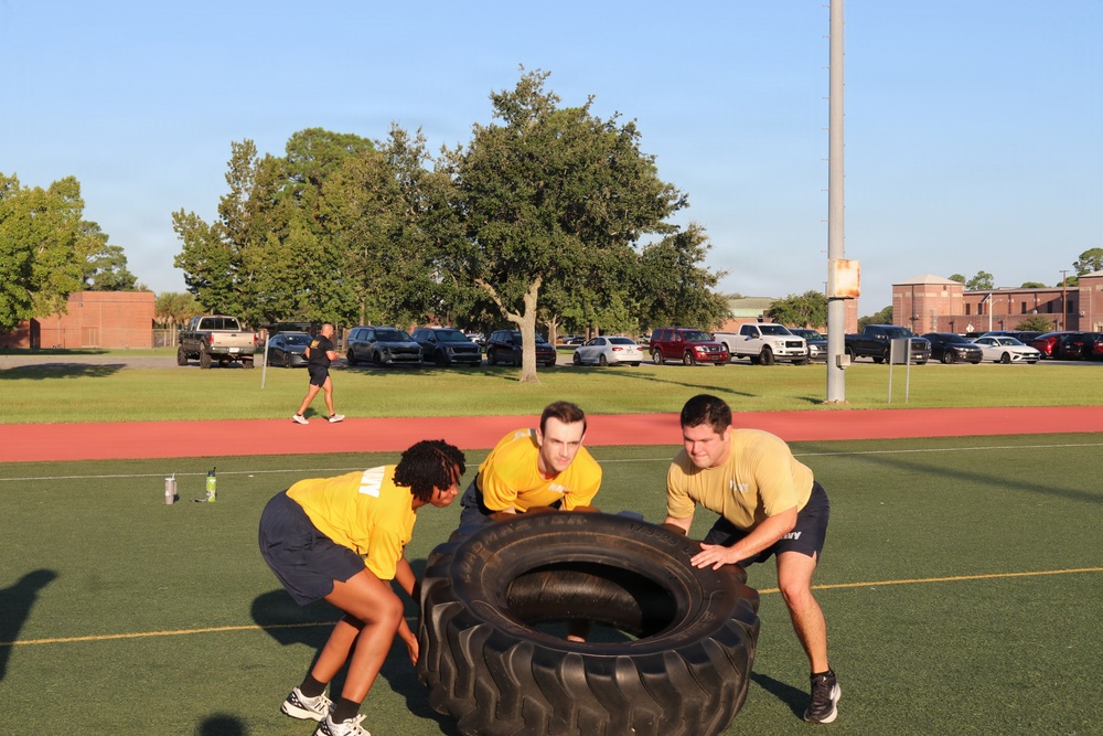 Three individuals wearing matching yellow shirts and dark shorts are performing a tire flip exercise on an artificial turf athletic field. The group appears to be engaged in a military or ROTC physical training session, evidenced by 'Army' text visible on their clothing. The setting is an outdoor sports complex with a running track, parking lot, and brick buildings visible in the background.