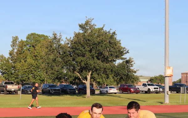 Three individuals wearing matching yellow shirts and dark shorts are performing a tire flip exercise on an artificial turf athletic field. The group appears to be engaged in a military or ROTC physical training session, evidenced by 'Army' text visible on their clothing. The setting is an outdoor sports complex with a running track, parking lot, and brick buildings visible in the background.