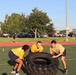 Three individuals wearing matching yellow shirts and dark shorts are performing a tire flip exercise on an artificial turf athletic field. The group appears to be engaged in a military or ROTC physical training session, evidenced by 'Army' text visible on their clothing. The setting is an outdoor sports complex with a running track, parking lot, and brick buildings visible in the background.