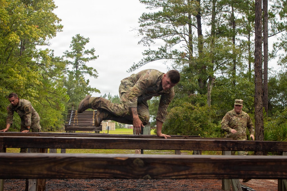 Three military personnel in camouflage uniforms are navigating a wooden obstacle course in an outdoor wooded setting. The central figure is actively climbing over a wooden beam obstacle, while two others are visible on either side. The setting appears to be a military training facility with pine trees and overcast skies in the background.
