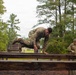 Three military personnel in camouflage uniforms are navigating a wooden obstacle course in an outdoor wooded setting. The central figure is actively climbing over a wooden beam obstacle, while two others are visible on either side. The setting appears to be a military training facility with pine trees and overcast skies in the background.