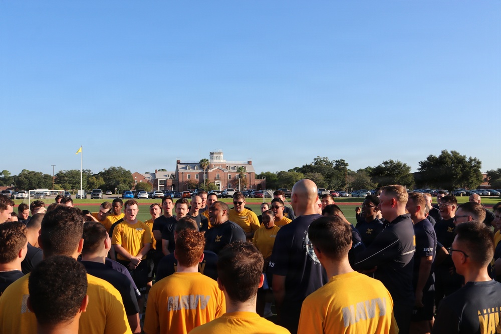 A large group of military personnel, likely U.S. Navy members based on visible 'NAVY' text on shirts, are gathered in a circle on an athletic field during what appears to be a morning formation or briefing. A bald individual stands centrally, appearing to address the group. The setting includes a red-brick institutional building, a flagpole with a yellow pennant, and a clear blue sky in the background.