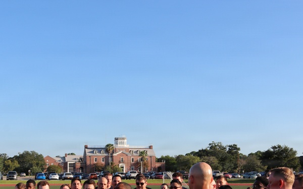 A large group of military personnel, likely U.S. Navy members based on visible 'NAVY' text on shirts, are gathered in a circle on an athletic field during what appears to be a morning formation or briefing. A bald individual stands centrally, appearing to address the group. The setting includes a red-brick institutional building, a flagpole with a yellow pennant, and a clear blue sky in the background.