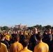 A large group of military personnel, likely U.S. Navy members based on visible 'NAVY' text on shirts, are gathered in a circle on an athletic field during what appears to be a morning formation or briefing. A bald individual stands centrally, appearing to address the group. The setting includes a red-brick institutional building, a flagpole with a yellow pennant, and a clear blue sky in the background.