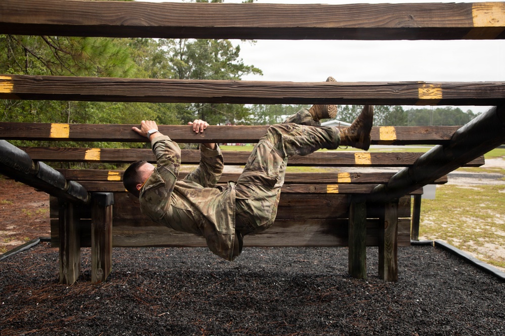 A soldier in camouflage uniform is navigating a military obstacle course, hanging and traversing underneath a series of horizontal wooden beams. The individual is suspended upside-down, gripping the beams with both hands while their legs are raised. The setting appears to be an outdoor military training facility with gravel ground cover and wooded surroundings.
