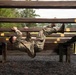 A soldier in camouflage uniform is navigating a military obstacle course, hanging and traversing underneath a series of horizontal wooden beams. The individual is suspended upside-down, gripping the beams with both hands while their legs are raised. The setting appears to be an outdoor military training facility with gravel ground cover and wooded surroundings.