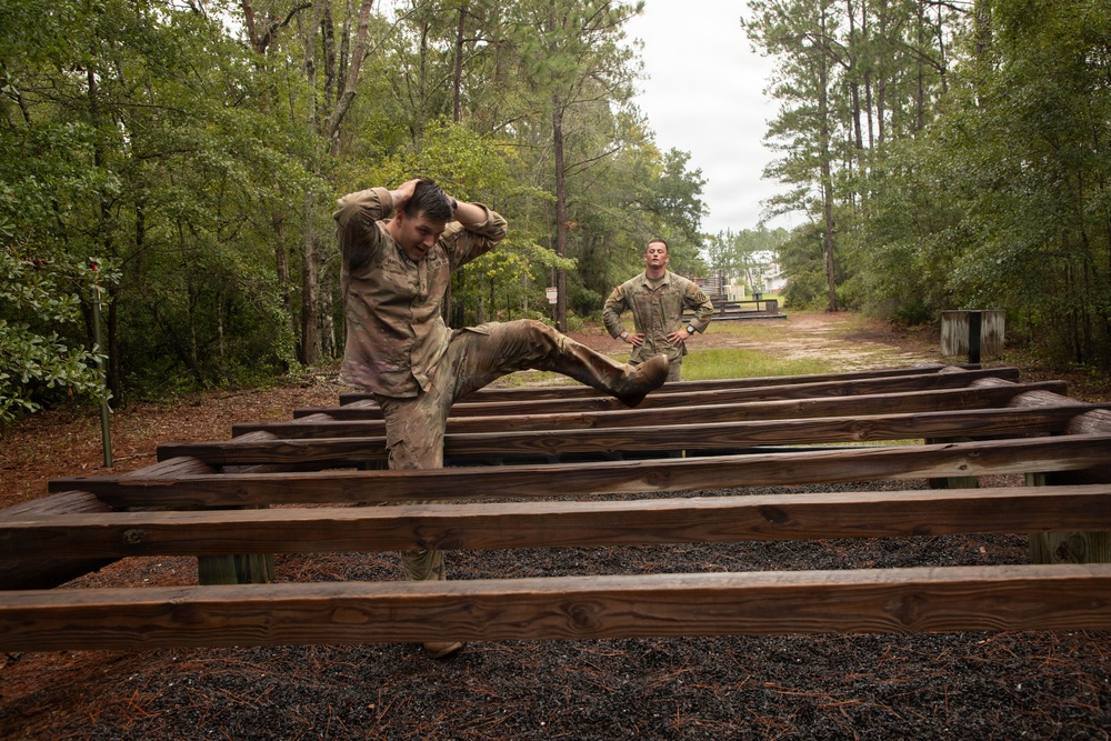 Two individuals in military camouflage uniforms are on what appears to be an outdoor obstacle course or physical training area. One soldier in the foreground is navigating a series of parallel wooden beams or rails, with hands behind their head and one leg raised, while a second soldier stands in the background observing with hands on hips. The setting is a wooded, overcast environment consistent with a military training facility.