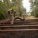 Two individuals in military camouflage uniforms are on what appears to be an outdoor obstacle course or physical training area. One soldier in the foreground is navigating a series of parallel wooden beams or rails, with hands behind their head and one leg raised, while a second soldier stands in the background observing with hands on hips. The setting is a wooded, overcast environment consistent with a military training facility.