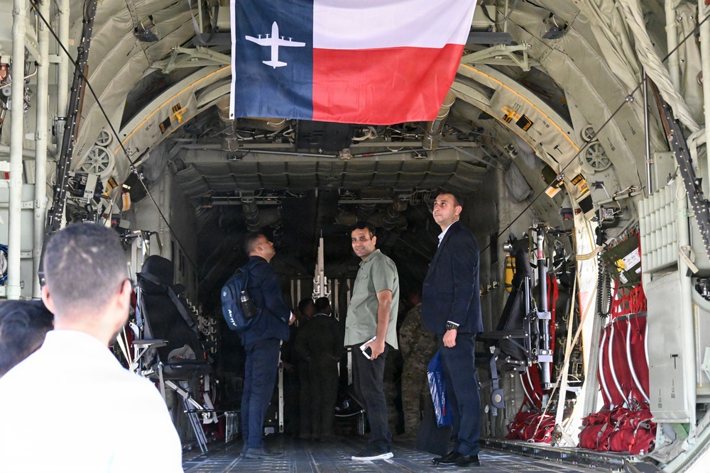 The image shows the interior of a large military cargo aircraft, likely a C-130 Hercules, with several civilians and at least one military personnel standing inside the open cargo bay. A modified Texas state flag featuring a C-130 aircraft silhouette in place of the lone star is prominently displayed hanging from the ceiling. The setting appears to be an airshow or public tour event based on the civilian attire and informal atmosphere.