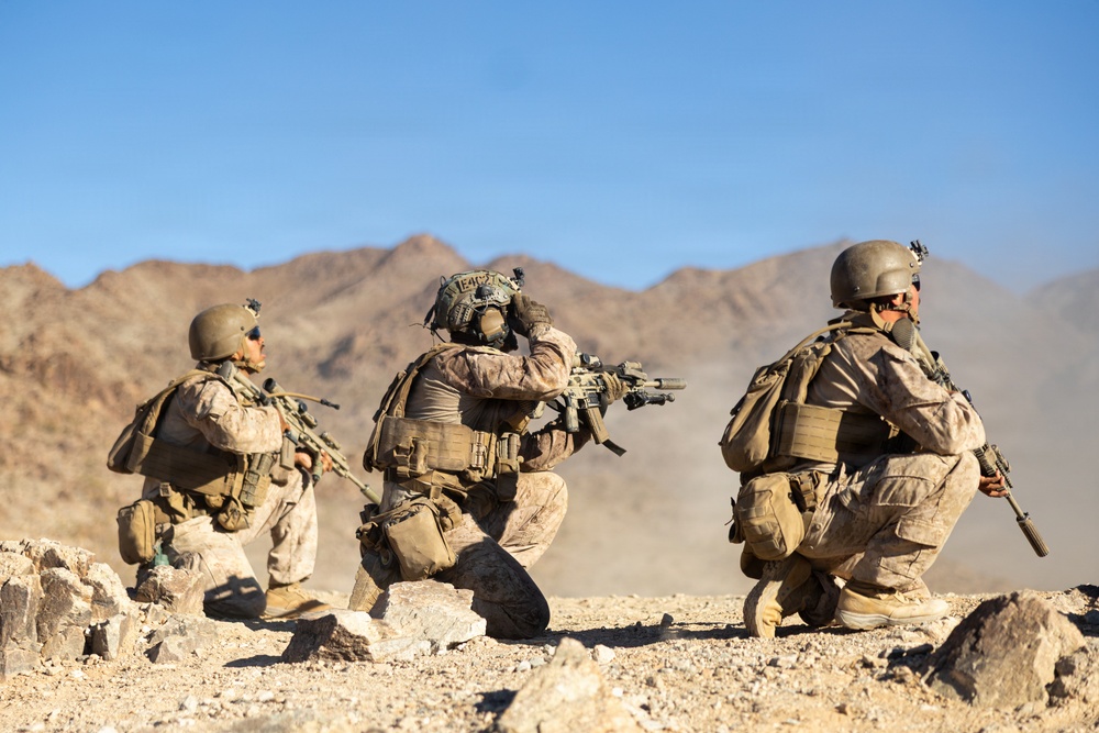 Three heavily equipped soldiers in desert camouflage uniforms are positioned in a kneeling/crouching formation on rocky, arid terrain. The center soldier is aiming a rifle forward while the others appear to be scanning their surroundings. The setting is a desert environment with rocky mountains visible in the background under a clear blue sky.