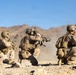Three heavily equipped soldiers in desert camouflage uniforms are positioned in a kneeling/crouching formation on rocky, arid terrain. The center soldier is aiming a rifle forward while the others appear to be scanning their surroundings. The setting is a desert environment with rocky mountains visible in the background under a clear blue sky.