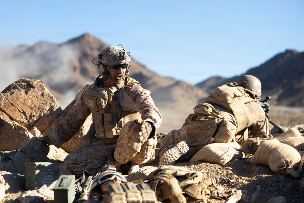 The image depicts military personnel in a desert rocky terrain, appearing to be engaged in a tactical field exercise or combat operation. At least three soldiers are visible, wearing desert camouflage uniforms and tactical gear, positioned among rocks and sandbags. The environment features arid mountainous terrain under a clear blue sky with dust or smoke visible in the background.