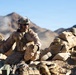 The image depicts military personnel in a desert rocky terrain, appearing to be engaged in a tactical field exercise or combat operation. At least three soldiers are visible, wearing desert camouflage uniforms and tactical gear, positioned among rocks and sandbags. The environment features arid mountainous terrain under a clear blue sky with dust or smoke visible in the background.