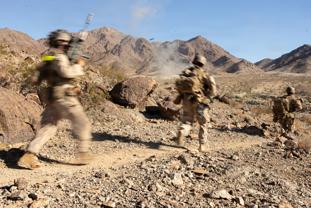 Three soldiers in desert camouflage uniforms and combat gear are moving rapidly across rocky, arid terrain toward a distant explosion or smoke cloud. The image captures motion blur on the figures, suggesting fast movement during what appears to be a military training exercise or combat operation. A mountainous desert landscape forms the backdrop under a clear blue sky.