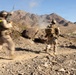 Three soldiers in desert camouflage uniforms and combat gear are moving rapidly across rocky, arid terrain toward a distant explosion or smoke cloud. The image captures motion blur on the figures, suggesting fast movement during what appears to be a military training exercise or combat operation. A mountainous desert landscape forms the backdrop under a clear blue sky.