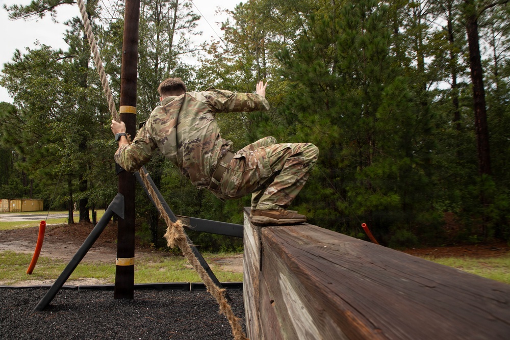 Two military personnel in camouflage uniforms are navigating an outdoor obstacle course, with one soldier climbing over a tall wooden wall while gripping a rope attached to a support pole. The setting appears to be a military training facility surrounded by pine trees, with rubber mulch ground cover beneath the obstacles. The image is captured from a low angle, emphasizing the height and physical challenge of the obstacle.