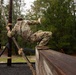 Two military personnel in camouflage uniforms are navigating an outdoor obstacle course, with one soldier climbing over a tall wooden wall while gripping a rope attached to a support pole. The setting appears to be a military training facility surrounded by pine trees, with rubber mulch ground cover beneath the obstacles. The image is captured from a low angle, emphasizing the height and physical challenge of the obstacle.