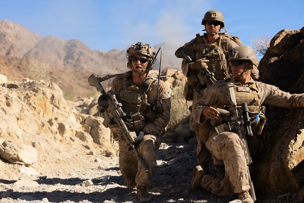 Three armed military personnel in full combat gear are positioned among rocky terrain in an arid, mountainous environment. They appear to be in a tactical formation, taking cover behind rocks while equipped with rifles, body armor, helmets, and communication equipment. The setting suggests a desert or semi-arid operational environment with rugged hills visible in the background.
