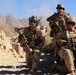 Three armed military personnel in full combat gear are positioned among rocky terrain in an arid, mountainous environment. They appear to be in a tactical formation, taking cover behind rocks while equipped with rifles, body armor, helmets, and communication equipment. The setting suggests a desert or semi-arid operational environment with rugged hills visible in the background.