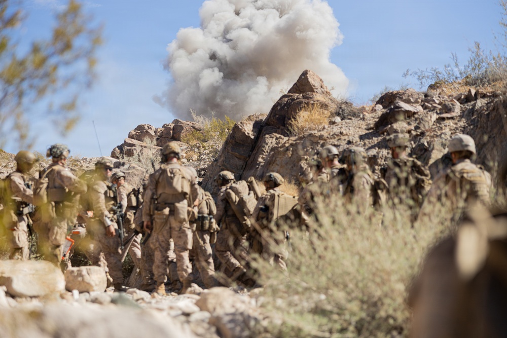 A group of military personnel in desert camouflage uniforms are moving through a rocky, arid terrain while a large smoke/dust cloud rises in the background from what appears to be an explosion or detonation. The soldiers are equipped with full combat gear including helmets, body armor, and backpacks. The scene appears to be either a combat operation or a military training exercise in a desert environment.
