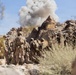 A group of military personnel in desert camouflage uniforms are moving through a rocky, arid terrain while a large smoke/dust cloud rises in the background from what appears to be an explosion or detonation. The soldiers are equipped with full combat gear including helmets, body armor, and backpacks. The scene appears to be either a combat operation or a military training exercise in a desert environment.
