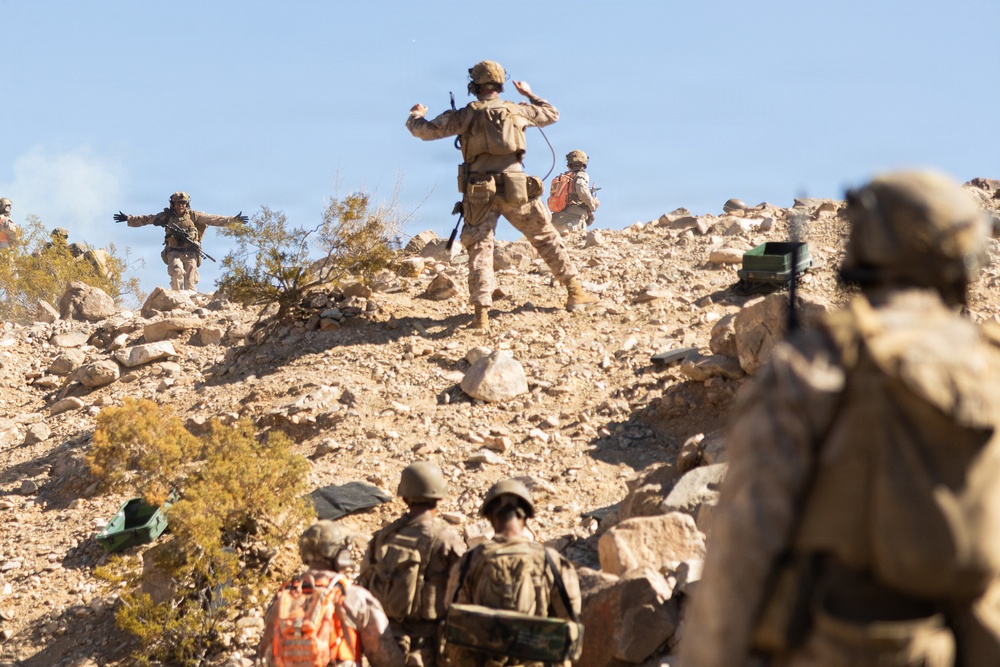 The image shows a group of military personnel in desert camouflage uniforms conducting what appears to be a training exercise or tactical operation on a rocky, arid hillside. Several soldiers are ascending the rocky terrain while others observe from below. One soldier near the top is making broad arm gestures, possibly signaling to others.
