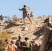 The image shows a group of military personnel in desert camouflage uniforms conducting what appears to be a training exercise or tactical operation on a rocky, arid hillside. Several soldiers are ascending the rocky terrain while others observe from below. One soldier near the top is making broad arm gestures, possibly signaling to others.