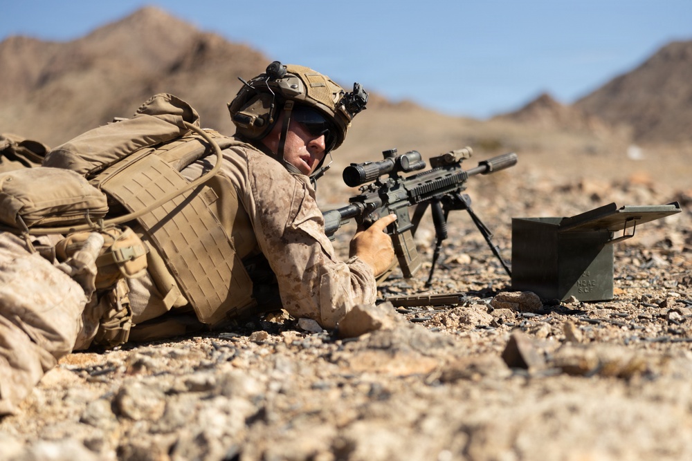 A military service member in desert camouflage (MARPAT pattern) is in a prone firing position in an arid, rocky terrain with mountains visible in the background. The soldier is aiming a scoped rifle mounted on a bipod, with an ammunition can visible nearby. The setting and equipment suggest a training exercise or operational deployment in a desert environment.