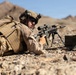A military service member in desert camouflage (MARPAT pattern) is in a prone firing position in an arid, rocky terrain with mountains visible in the background. The soldier is aiming a scoped rifle mounted on a bipod, with an ammunition can visible nearby. The setting and equipment suggest a training exercise or operational deployment in a desert environment.