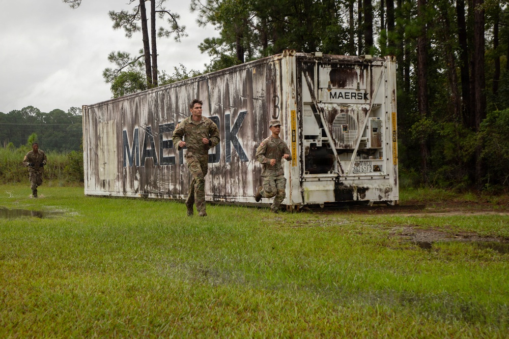 Three military personnel in camouflage uniforms are running past a weathered Maersk shipping container in a wooded, wet outdoor environment. The scene appears to be a training exercise or physical fitness activity conducted in overcast, rainy conditions. The ground shows standing water and saturated grass, suggesting recent or ongoing rainfall.