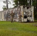 Three military personnel in camouflage uniforms are running past a weathered Maersk shipping container in a wooded, wet outdoor environment. The scene appears to be a training exercise or physical fitness activity conducted in overcast, rainy conditions. The ground shows standing water and saturated grass, suggesting recent or ongoing rainfall.