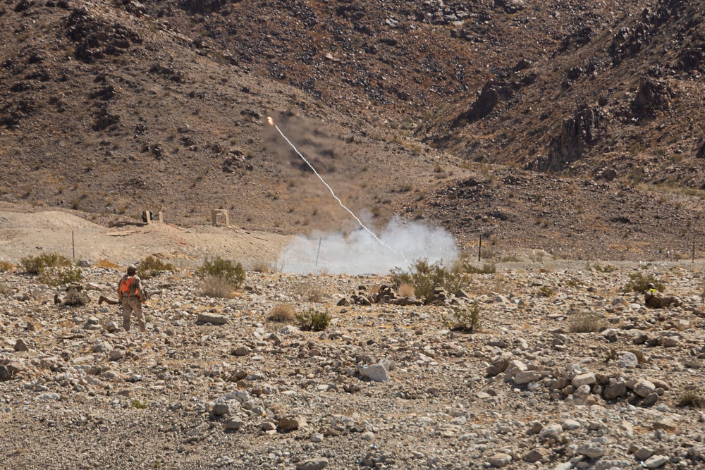 A person wearing an orange safety vest stands in a rocky desert terrain while a projectile or flare is visibly in flight, leaving a white smoke trail against a mountainous backdrop. The scene appears to be a controlled test, demonstration, or training exercise in an arid, remote location. Smoke is visible near the launch point on the ground.