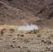 A person wearing an orange safety vest stands in a rocky desert terrain while a projectile or flare is visibly in flight, leaving a white smoke trail against a mountainous backdrop. The scene appears to be a controlled test, demonstration, or training exercise in an arid, remote location. Smoke is visible near the launch point on the ground.