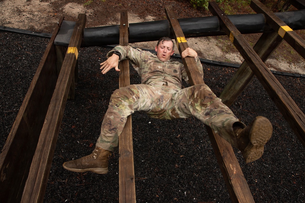 A soldier in OCP (Operational Camouflage Pattern) uniform is navigating a military obstacle course, appearing to crawl or slide under a low horizontal barrier supported by wooden rails. The individual is lying on their back on a rubber mulch surface, gripping the wooden rails while maneuvering beneath the obstacle. The soldier appears physically exerted, with mouth open and eyes closed.
