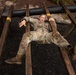 A soldier in OCP (Operational Camouflage Pattern) uniform is navigating a military obstacle course, appearing to crawl or slide under a low horizontal barrier supported by wooden rails. The individual is lying on their back on a rubber mulch surface, gripping the wooden rails while maneuvering beneath the obstacle. The soldier appears physically exerted, with mouth open and eyes closed.