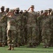 A large formation of U.S. Army soldiers in Operational Camouflage Pattern (OCP) uniforms are standing at attention and rendering a salute during a formal military ceremony. The soldiers are arranged in multiple rows on a grass field, suggesting a parade, review, or official event. A female soldier in the foreground appears to be a senior NCO based on visible rank insignia and the 101st Airborne Division patch on her shoulder.