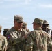 A group of U.S. Army soldiers in OCP (Operational Camouflage Pattern) uniforms are gathered outdoors, with a senior soldier appearing to pin an award or badge onto another soldier's uniform. Multiple soldiers are wearing the 82nd Airborne Division patch on their left shoulders. The setting appears to be an outdoor military ceremony or formation.