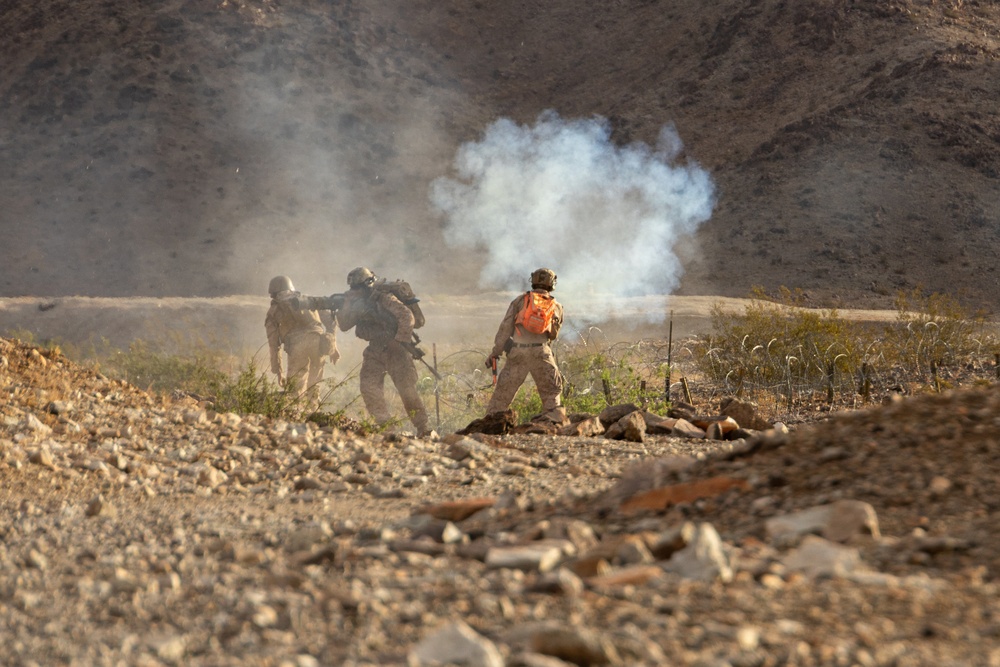 The image shows what appears to be military personnel in a desert/arid terrain environment with smoke visible in the background. Three individuals in military-style camouflage uniforms are visible, with one wearing a distinctive orange backpack. The scene suggests a training exercise or field operation in a rocky, mountainous desert landscape.