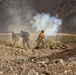 The image shows what appears to be military personnel in a desert/arid terrain environment with smoke visible in the background. Three individuals in military-style camouflage uniforms are visible, with one wearing a distinctive orange backpack. The scene suggests a training exercise or field operation in a rocky, mountainous desert landscape.