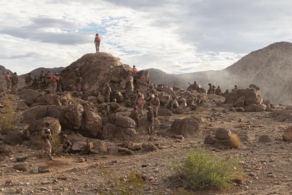A large group of military personnel in desert camouflage uniforms are dispersed across a rocky, arid terrain with mountains in the background. Several individuals are wearing bright orange vests, distinguishing them from others, suggesting a training exercise with observer/controller or opposing force roles. The scene appears to be a military field training exercise in a desert environment.