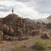 A large group of military personnel in desert camouflage uniforms are dispersed across a rocky, arid terrain with mountains in the background. Several individuals are wearing bright orange vests, distinguishing them from others, suggesting a training exercise with observer/controller or opposing force roles. The scene appears to be a military field training exercise in a desert environment.