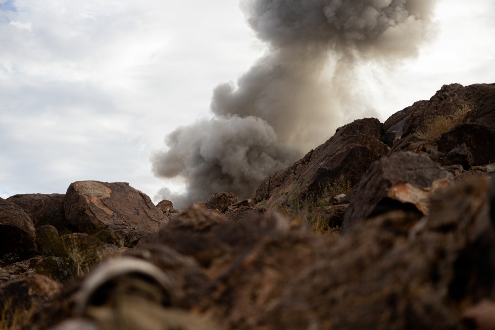 A ground-level photograph captures a large plume of dark gray smoke rising above a rocky, arid terrain. The camera angle is low, positioned among reddish-brown boulders and rocks, giving a dramatic perspective of the smoke column ascending into an overcast sky. The scene suggests an explosion, controlled detonation, or significant fire occurring just beyond the rock formation.
