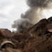 A ground-level photograph captures a large plume of dark gray smoke rising above a rocky, arid terrain. The camera angle is low, positioned among reddish-brown boulders and rocks, giving a dramatic perspective of the smoke column ascending into an overcast sky. The scene suggests an explosion, controlled detonation, or significant fire occurring just beyond the rock formation.