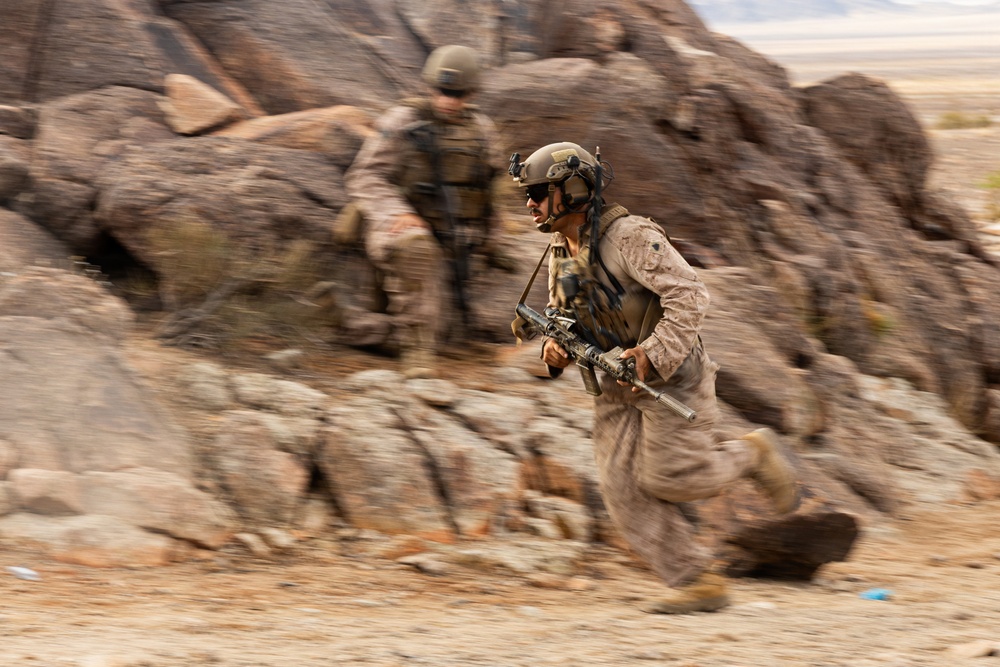 Two military personnel in desert camouflage uniforms are moving rapidly through a rocky, arid terrain. The foreground soldier is in sharp focus while running and carrying a rifle, with motion blur indicating fast movement. The environment appears to be a desert training area or operational zone with large rock formations.