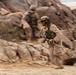 Two military personnel in desert camouflage uniforms are moving rapidly through a rocky, arid terrain. The foreground soldier is in sharp focus while running and carrying a rifle, with motion blur indicating fast movement. The environment appears to be a desert training area or operational zone with large rock formations.