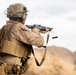 A military service member in desert MARPAT camouflage is shown from behind, aiming a shotgun in an arid, desert environment. The soldier is wearing full combat gear including a tactical vest, helmet with communications equipment, and a name tape reading 'DAVIDGE'. The background features rocky desert terrain and an overcast sky.