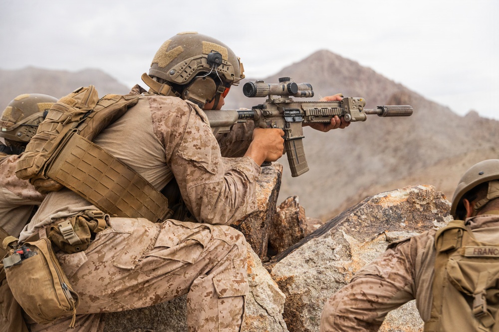 The image shows military personnel in desert camouflage uniforms taking up a firing position behind rocky cover in an arid, mountainous environment. The primary subject is aiming a rifle equipped with an optical scope and suppressor over a rock formation. At least two additional personnel are visible in the frame.