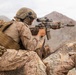 The image shows military personnel in desert camouflage uniforms taking up a firing position behind rocky cover in an arid, mountainous environment. The primary subject is aiming a rifle equipped with an optical scope and suppressor over a rock formation. At least two additional personnel are visible in the frame.