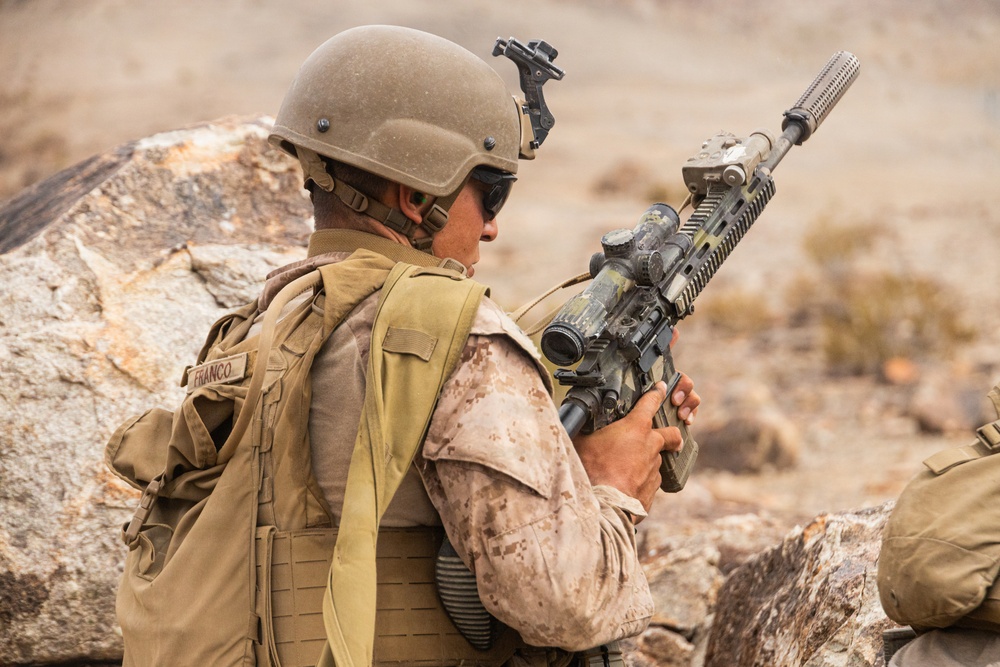 A soldier wearing desert camouflage uniform and tactical gear is positioned behind rocky cover in an arid environment. The individual, identified by a name tape reading 'FRANCO', is holding an assault rifle equipped with a scope and suppressor. The setting and equipment suggest a military training exercise or operational deployment in a desert terrain.