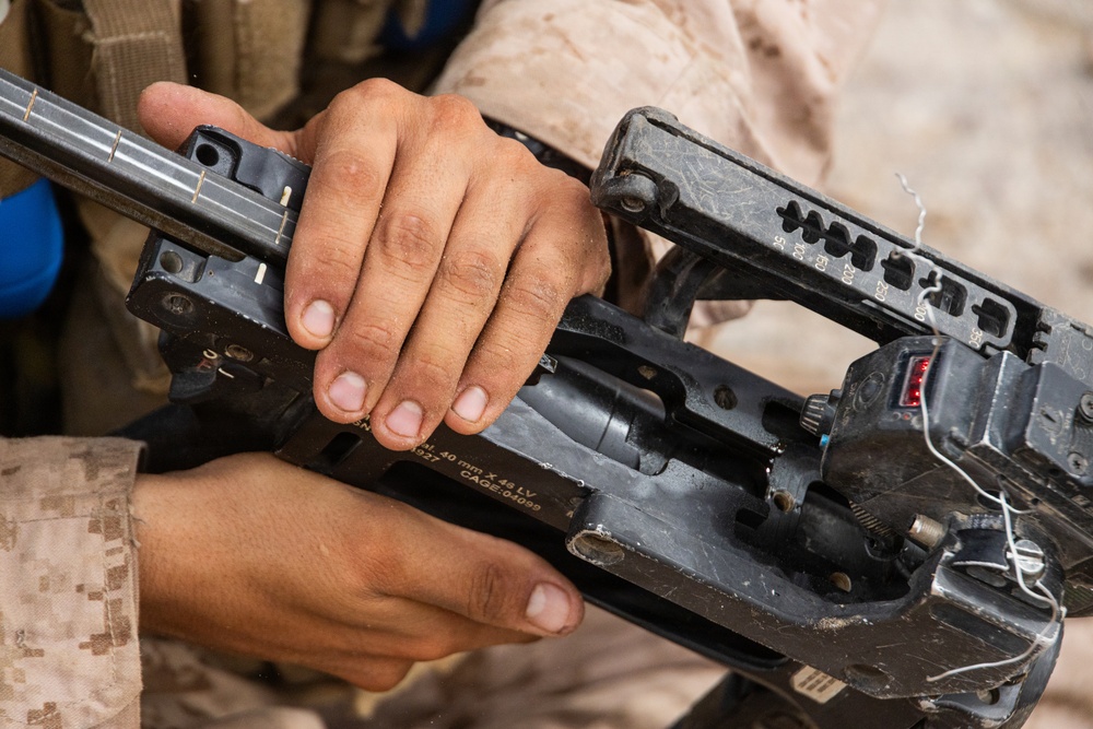 A person wearing MARPAT desert camouflage uniform is handling a disassembled or open grenade launcher, likely an M320 or similar 40mm grenade launcher. The weapon is open/broken down, exposing internal components. Visible markings on the weapon reference '40mm x 46 LV' ammunition caliber and a CAGE code.